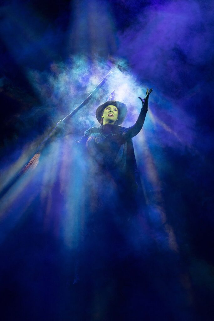 Sheridan Adams as Elphaba in Wicked holding her broomstick and shows the illusion of flying in the dark sky, singing Defying Gravity. 